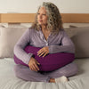 Woman in lavender sleepwear sitting on a bed, holding a Purple Yogibo Mega Moon Pillow across her lap and hugging it under her arms.