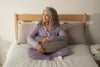 Woman in lavender sleepwear sitting on a bed, holding a grey Yogibo Mega Moon Pillow across her lap and hugging it under her arms.