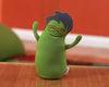 A green, travel-sized stuffed toy resembling an egg with arms and glasses, set against a blurred background.